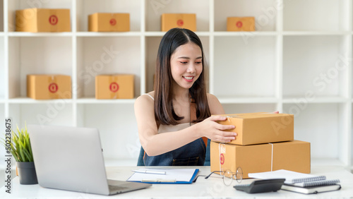 Happy young Asian business owner woman prepare parcel box and check online orders of product for deliver to customer on laptop computer. Shopping Online concept. © amnaj