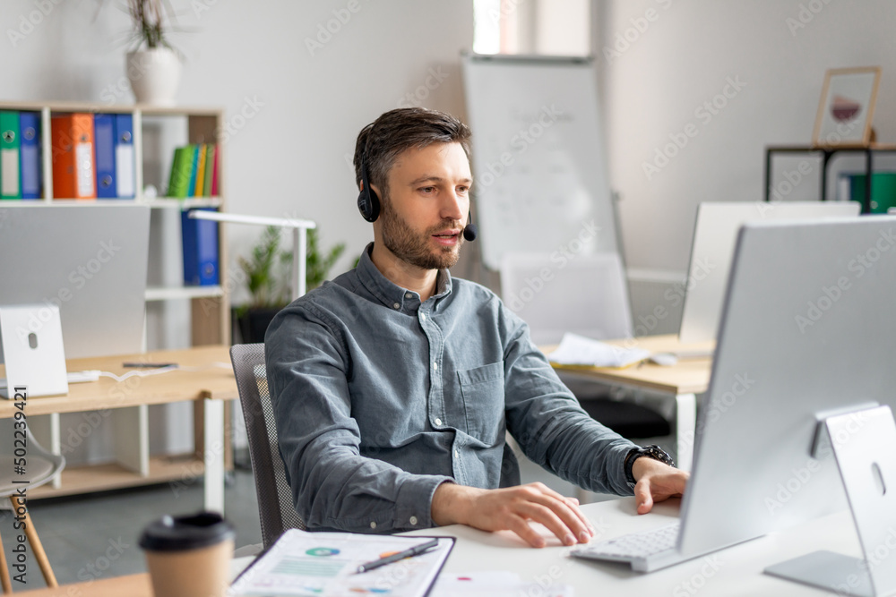 Hotline support service. Handsome mature male call center operator in headset working on modern computer in office Hotline support service. Handsome mature male call center operator in headset working on modern computer in office