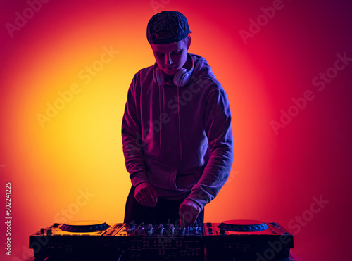 Portrait of young man making sounds with professional dj mixer isolated over gradient red yellow background in neon light