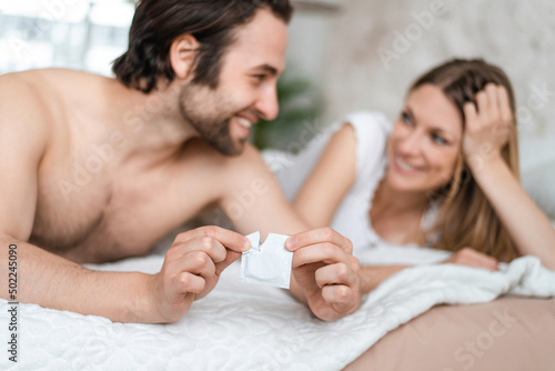 Happy young couple lying in bed and opening condom, selective focus. Pregnancy and STD control concept