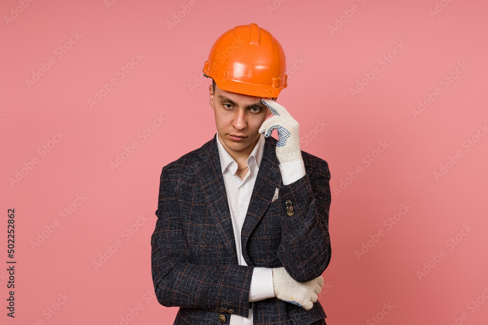 young male engineer in a safety helmet and jacket thinking on a pink background