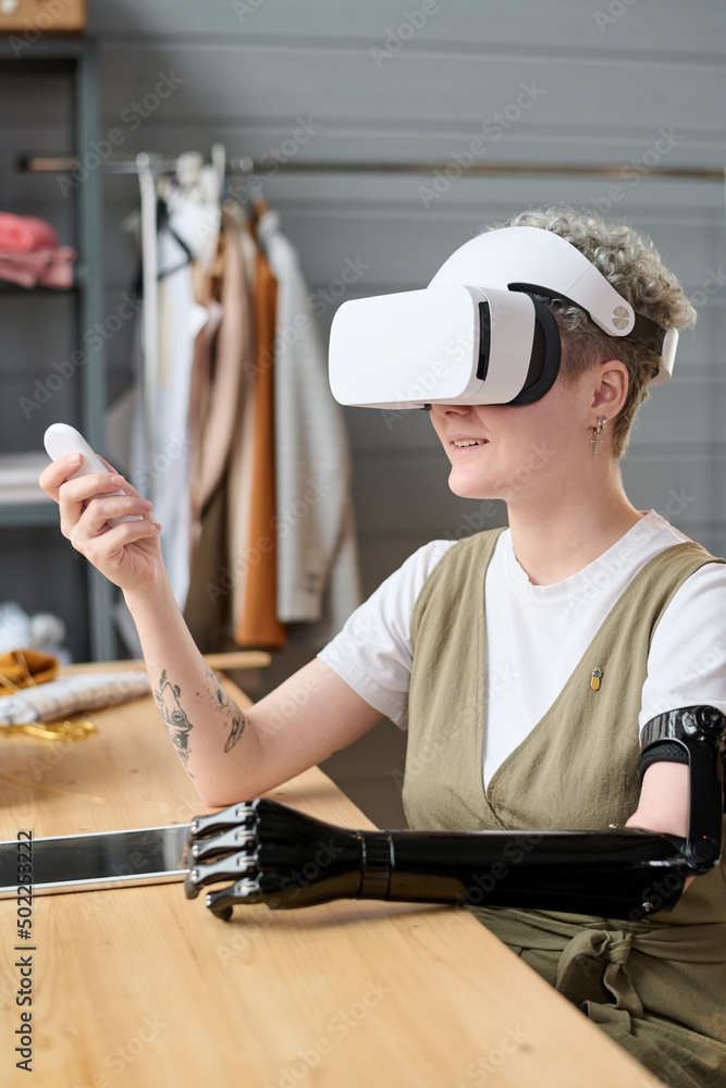 Young modern woman with myoelectric arm in vr headset using remote control while making or preparing presentation of new project Young modern woman with myoelectric arm in vr headset using remote control while making or preparing presentation of new project