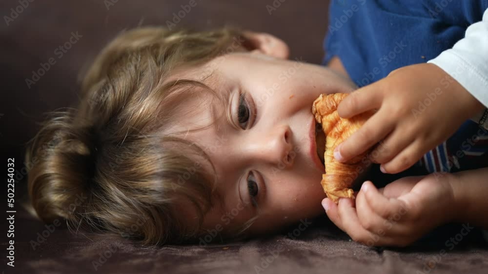 Child eating croissant one little boy lying on sofa eats carb food Child eating croissant one little boy lying on sofa eats carb food