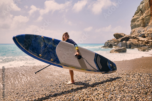 Young paddle female surfer stands on a beach. Stand up paddle boarding. Summer outdoor recreational activities. 