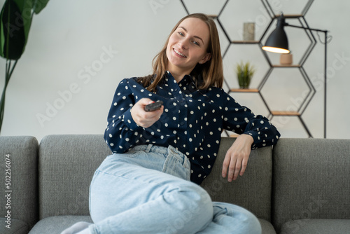 Young cheerful woman is watching a funny movie on cable TV, switching channels using a remote control at home. © Grustock