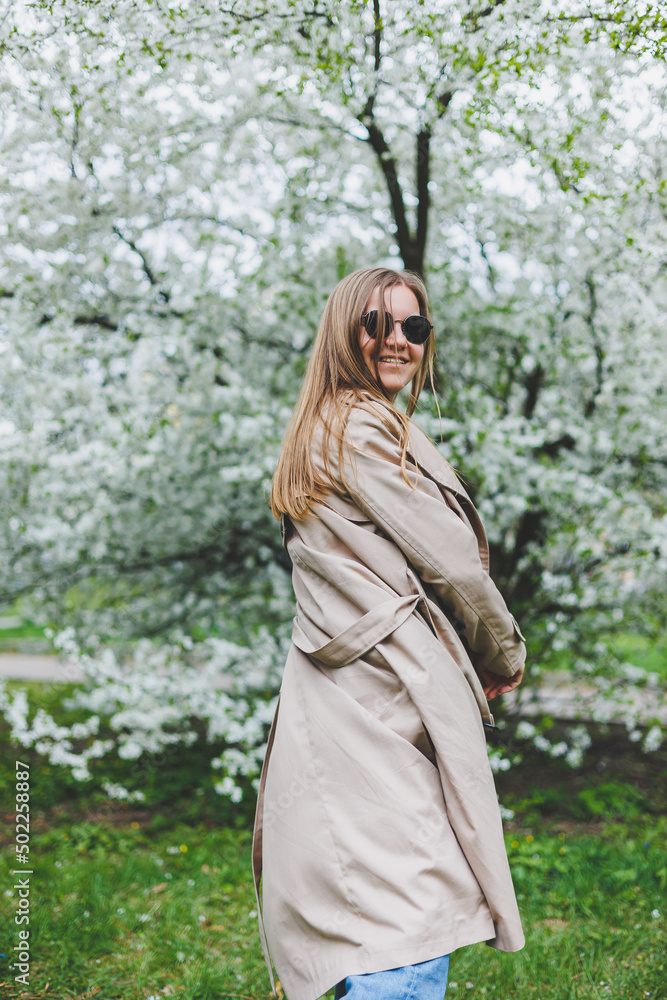 A young woman with blond hair enjoys a blooming spring garden. Travel, spring break. Fashionable style. A woman in sunglasses and a beige trench coat runs through a flowering park. Selection focus A young woman with blond hair enjoys a blooming spring garden. Travel, spring break. Fashionable style. A woman in sunglasses and a beige trench coat runs through a flowering park. Selection focus
