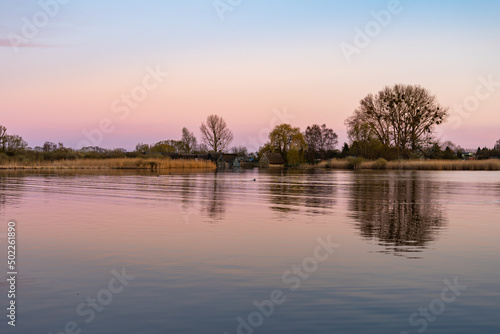 Small houses next to a lake. Wooden buildings next to reed plants. Water is is reflecting the rural landscape. Old architecture and bare trees in the environment. © 1take1shot