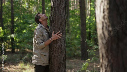 Side view Caucasian man standing at tree trunk looking up in woodland. Male traveler enjoying calm spring summer day in forest outdoors hugging plant. Connection with nature and tranquility photo