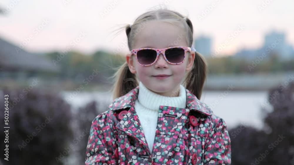 Portrait of a fashionable cute Caucasian child girl who is wearing stylish sunglasses. A little girl stands alone in a city public park, looks at the camera and smiles. Fashion and lifestyle concept Portrait of a fashionable cute Caucasian child girl who is wearing stylish sunglasses. A little girl stands alone in a city public park, looks at the camera and smiles. Fashion and lifestyle concept