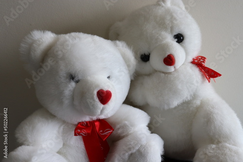 Little white teddy bears with red bow on neck, red heart nose, black button eyes, cuddling bears. white wall background