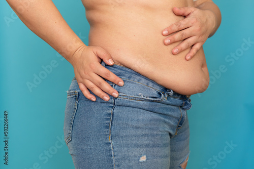 Side view of unrecognizable fat plump obese plus-size overweight woman stand in blue jeans, putting hand on excess naked belly on blue background. Body positive, obesity, weight loss, liposuction.