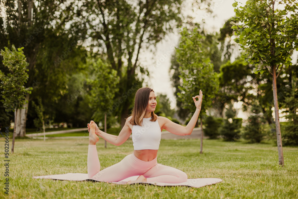 Young fit woman practice yoga on gymnastic mat in green park on grass. Stand in dove pose, do stretching exercises keeping balance in sportswear. Concept of healthy lifestyle and harmony with body.