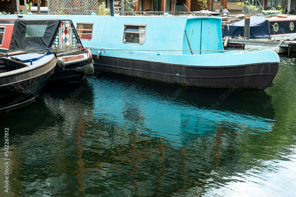 Boats along a London canal that are reflected in the water of the river, dominance of blue and green colors.