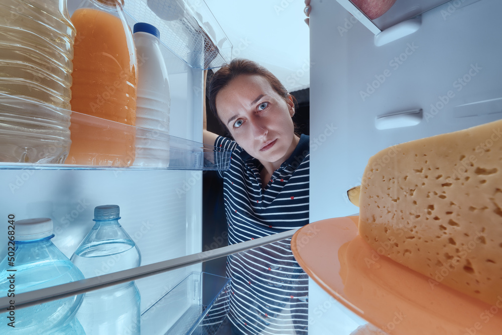 Serious girl looks thoughtfully into fridge full of different food