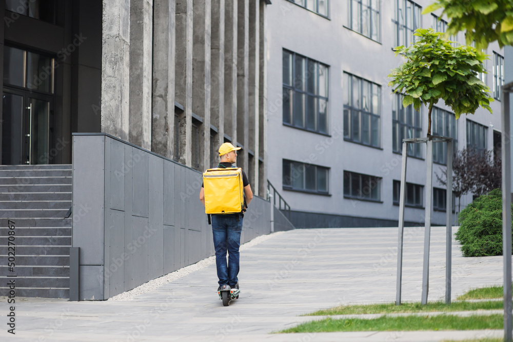 Young man with an yellow backpack-satchel on his back in casual summer clothes is moving along the pedestrian sidewalk on an electric scooter from left to right in the city