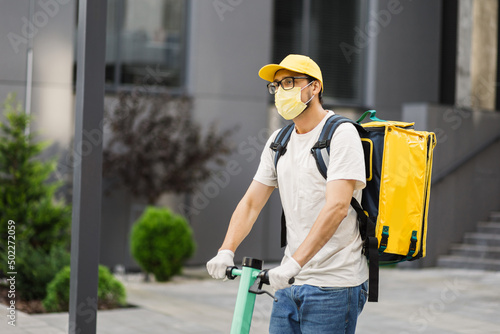 Courier deliver food on electric scooter wearing yellow facial protective mask, working during covid-19 pandemic. Boy delivering hot food to costumers. Delivery service from cafes and restaurants.