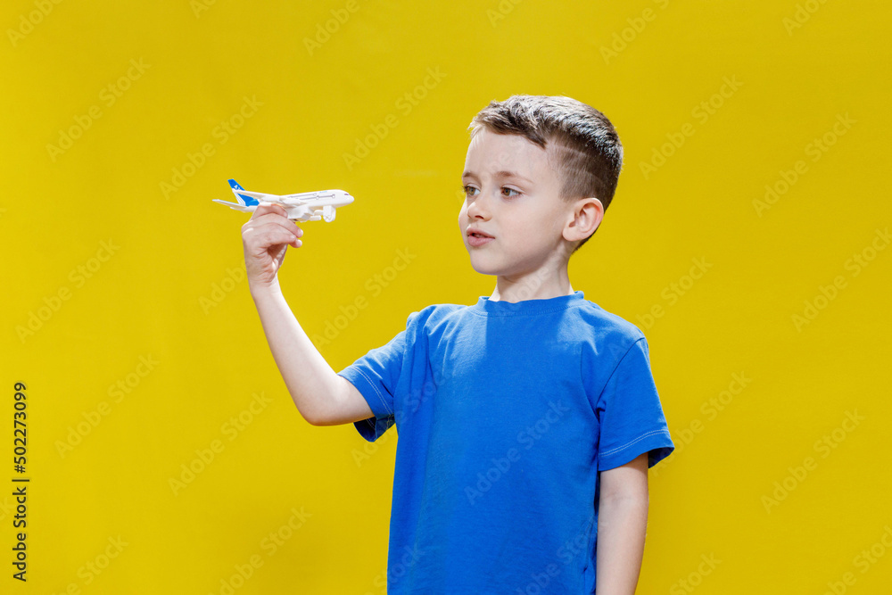 Little boy in a blue T-shirt holds a toy white plane and plays on a yellow background. Copy space. Travel concept