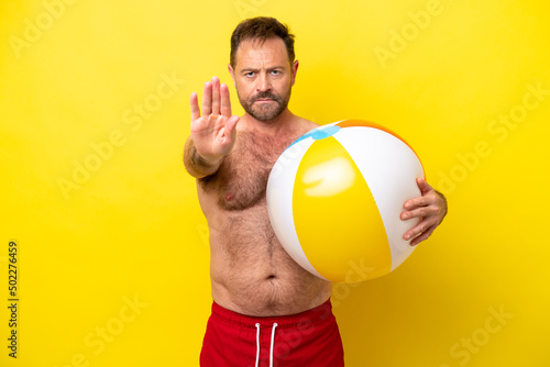 Middle age caucasian man holding beach ball isolated on yellow background making stop gesture