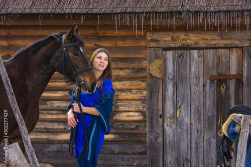 A beautiful young girl holds a horse by the bridle, against the background of a log wall