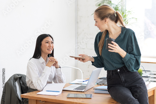 Two young women in the office. Talking, smiling, coffe break, pause. Diversity, asian female. Coffee break, chat and small talk with friends and colleagues, team members photo