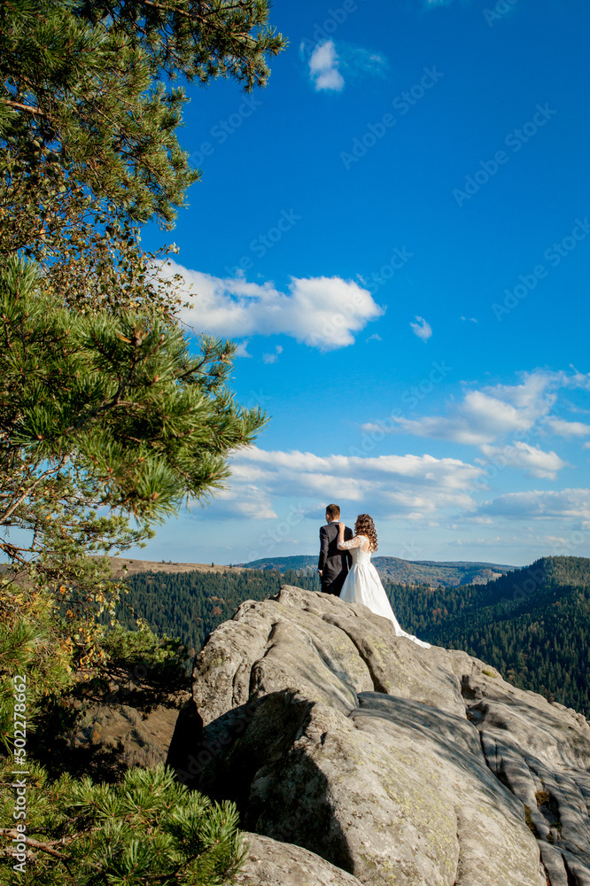 Beautiful newlyweds hugging against the backdrop of rocks and mountains. Stylish bride and beautiful bride are standing on the cliff. Wedding portrait. Family photo