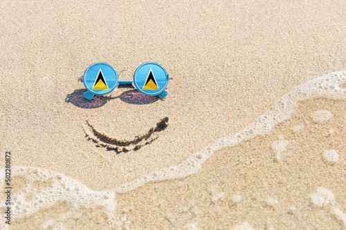 A painted smile on the sand and sunglasses with the flag of the Saint Lucia. The concept of a positive and successful holiday in the resort of the Saint Lucia.