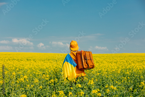 Woman in yellow hoodie with ukrainian flag and suitcase in rapeseed field