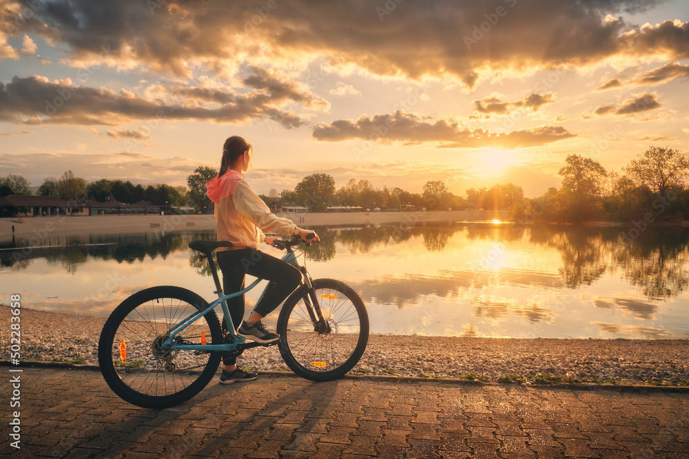 Fototapeta premium Woman riding a mountain bike near lake at sunset in summer. Colorful landscape with sporty girl riding a bicycle, beach, beautiful sky reflected in water in park in spring. Sport and travel. Biking