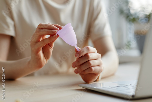 Closeup of young woman holding pink menstrual cup by laptop and researching feminine products photo