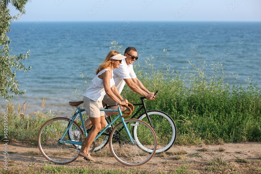 Mature couple riding bicycles along sea shore on summer day