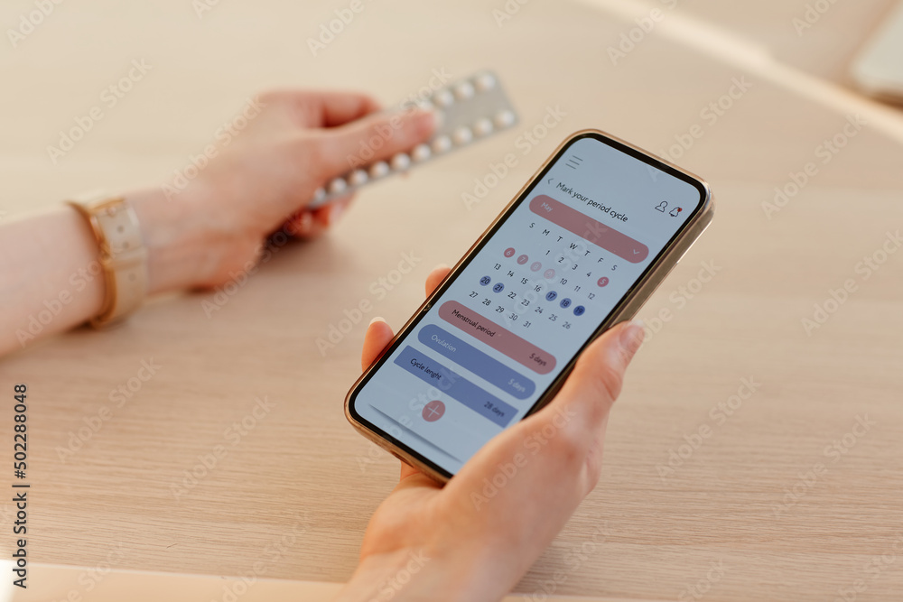 Close up of young woman calculating menstrual cycle using mobile app and holding birth control pills - obrazy, fototapety, plakaty 