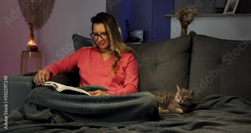 Mulher jovem lendo um livro sentada no sofá ao lado de um gato 