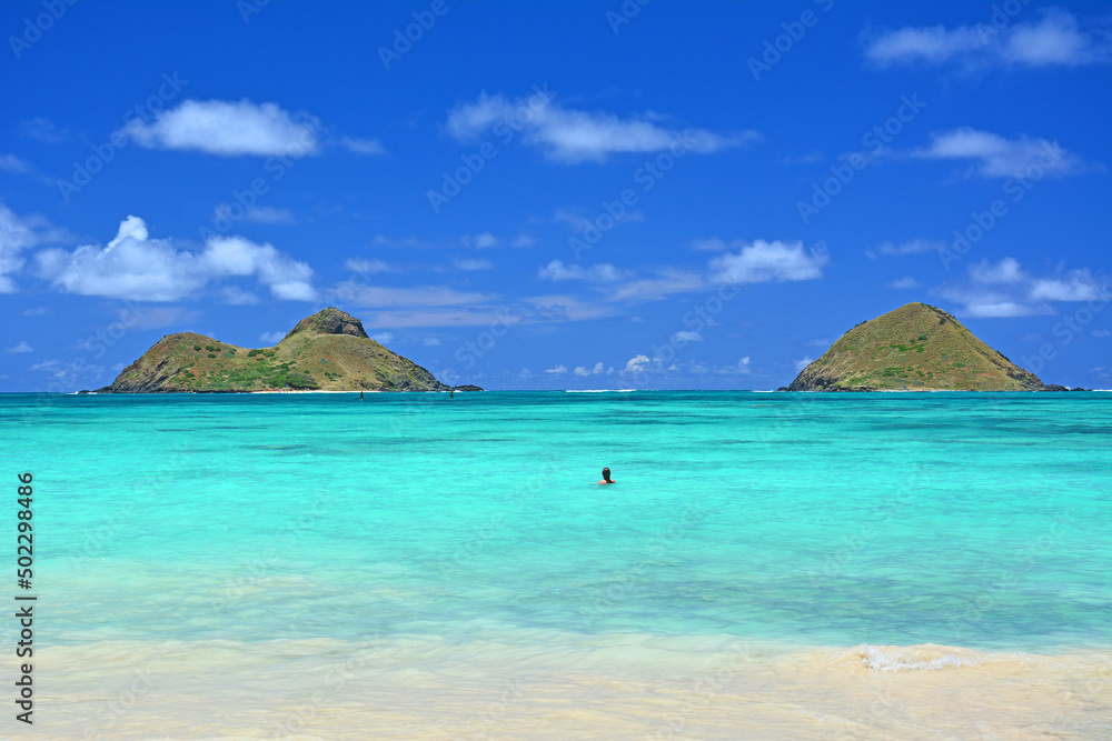 A lone swimmer enjoying turquoise waters and a lovely view of the Mokes aka Na Mokulua on a clear sunny day at Lanikai Beach on the windward side of Oahu, Hawaii. 