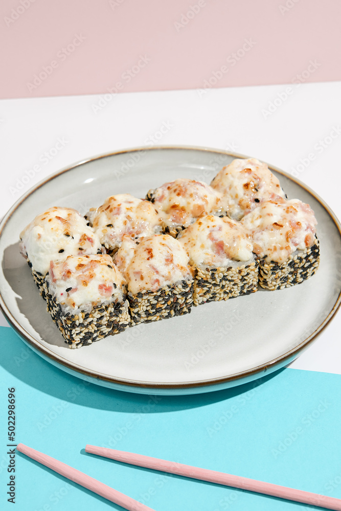 Baked Maki roll with bacon on ceramic plate. Hot sushi roll with sesame outside, bacon and cheese topped on coloured background. Japanese menu concept. Hot maki sushi in modern style. Baked Maki roll with bacon on ceramic plate. Hot sushi roll with sesame outside, bacon and cheese topped on coloured background. Japanese menu concept. Hot maki sushi in modern style.
