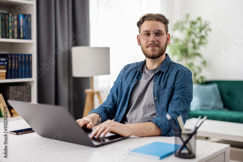 Wallpaper Mural Portrait of positive male freelancer sitting at home office with modern laptop on table. Young bearded guy wearing eyeglasses and casual denim shirt. Torontodigital.ca