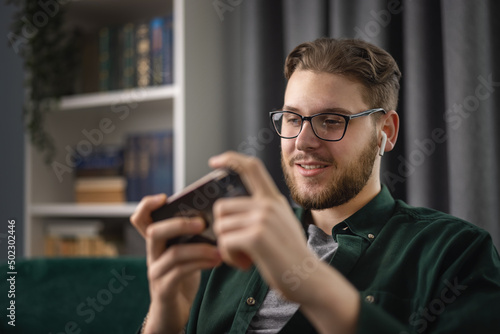 Happy young man in wireless earphones sitting on couch and playing video games on smartphone. Relaxed caucasian guy using modern gadgets for taking fun at cozy apartment.