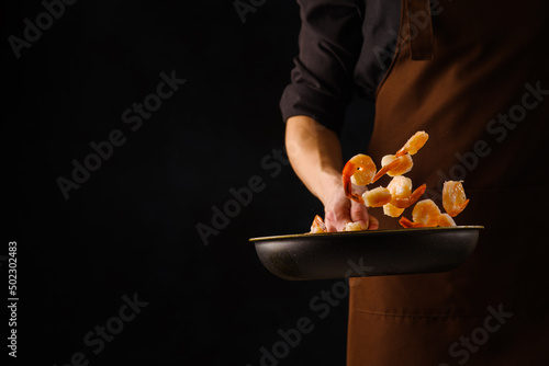 Professional chef cooks shrimp in a frying pan. Seafood cooking, healthy vegetarian food. Black background, frozen food in flight. Restaurant, hotel, banquet, picnic, recipe book.