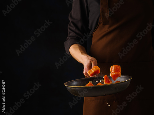 A professional chef prepares pieces of red fish - tuna, salmon, trout in a pan on a black background. Levitation. The concept is recipes for fish and seafood dishes. Restaurant, hotel, banner.