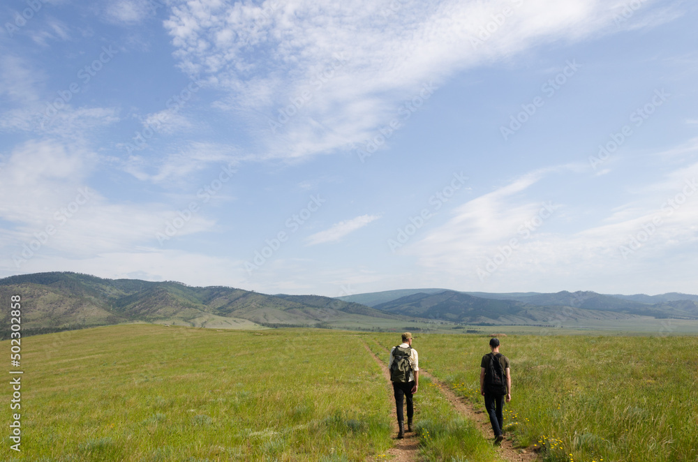 Two travelers walk along the road among the green hills. Hiking in nature. Two travelers walk along the road among the green hills. Hiking in nature.