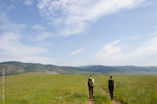 Two travelers walk along the road among the green hills. Hiking in nature.