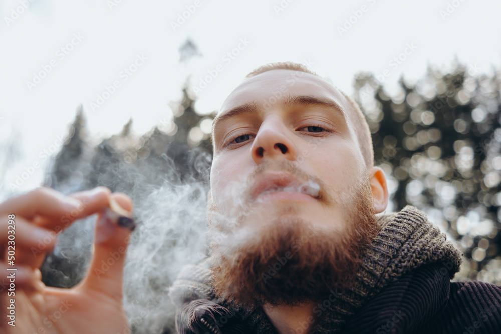 Young man beard smokes cigarette or tobacco. Guy is addicted to cigarettes and always smokes lot of tobacco every day. He smokes cigarette outdoors on background of lake in park. Cool guy blows smoke Young man beard smokes cigarette or tobacco. Guy is addicted to cigarettes and always smokes lot of tobacco every day. He smokes cigarette outdoors on background of lake in park. Cool guy blows smoke