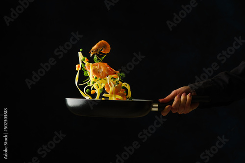 The process of cooking italian pasta with seafood and vegetables in a frying pan on a black background by a professional chef. Levitation. Recipes for healthy vegetarian food with seafood.