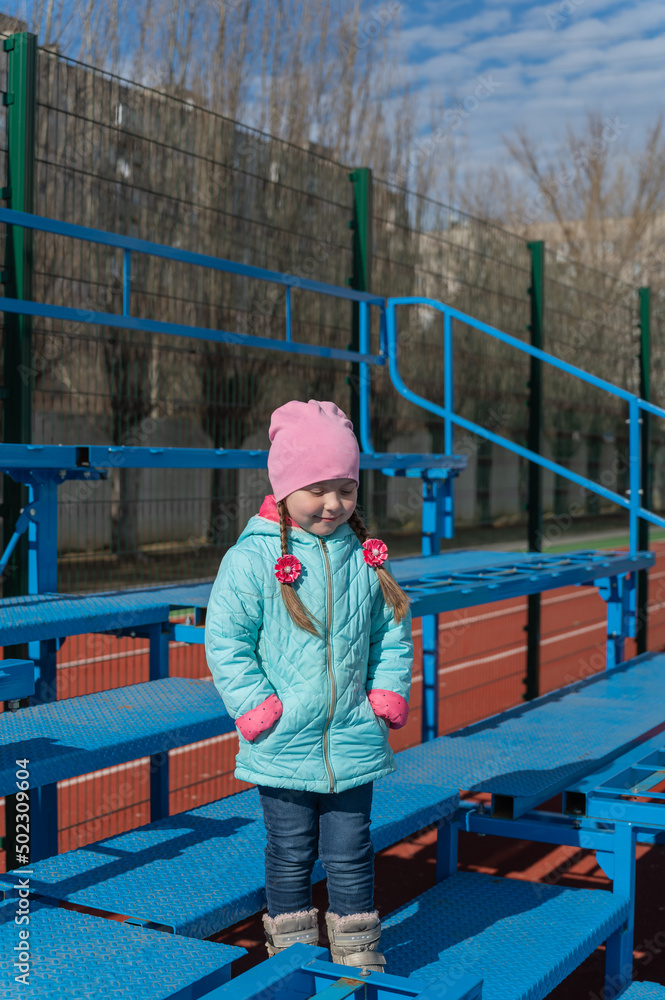 Full-length portrait of a five-year-old girl in an empty sports stand. The child stands on the blue tribune of the school stadium holding her hands in her jacket pockets. Full-length portrait of a five-year-old girl in an empty sports stand. The child stands on the blue tribune of the school stadium holding her hands in her jacket pockets.