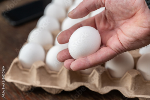 A man is holding a chicken egg in front of an egg tray. Adult male holding a white raw egg. Open recycled tray. Selective Focus.