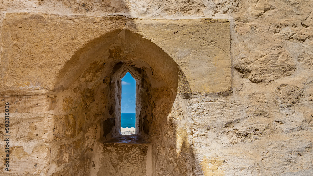 A fragment of the ancient Citadel of Qaitbay. A thick stone wall with a rough surface. Through a narrow window- loophole you can see the blue sky, the sea. Egypt. Alexandria