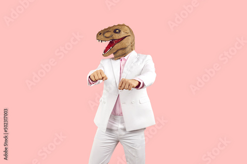 Weird guy in funny disguise dancing against pastel pink studio background. Cheerful eccentric man in white suit and silly ugly wacky masquerade dinosaur mask having fun at crazy party