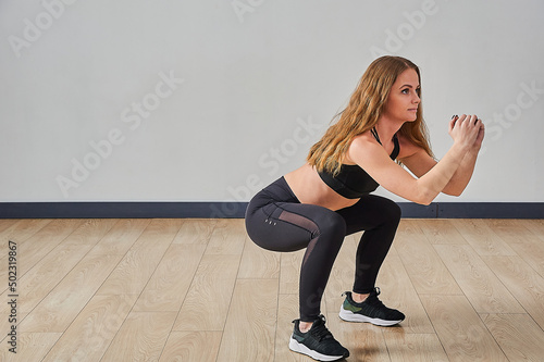 A young beautiful woman in a tracksuit holding her hands in front of her doing squats in the gym, copy space