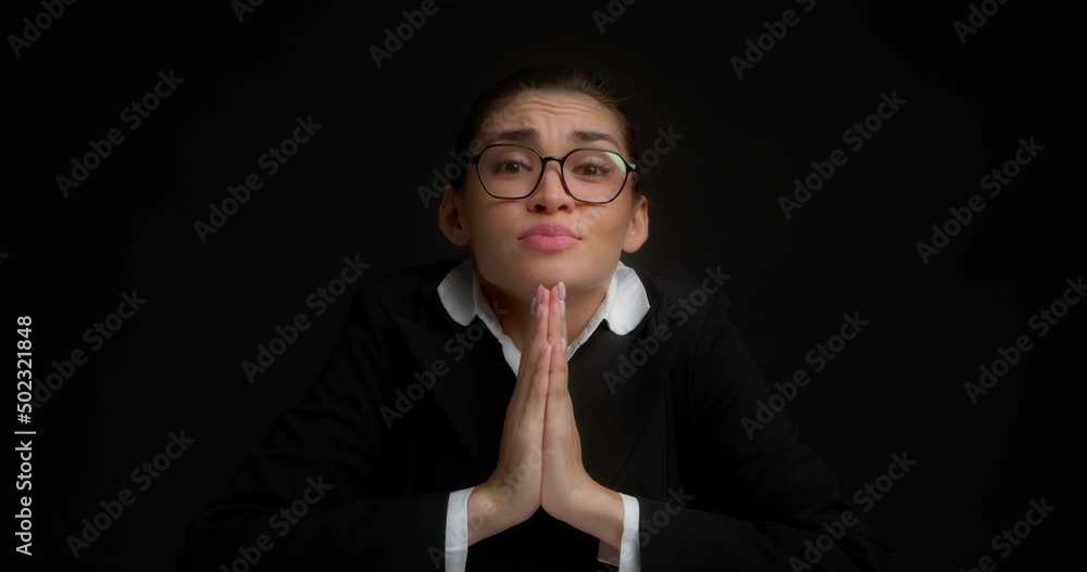Pretty brunette in glasses and business clothes stands on an isolated black background and pleadingly looks into the camera, asks for something, holds her palms together. Pretty brunette in glasses and business clothes stands on an isolated black background and pleadingly looks into the camera, asks for something, holds her palms together.