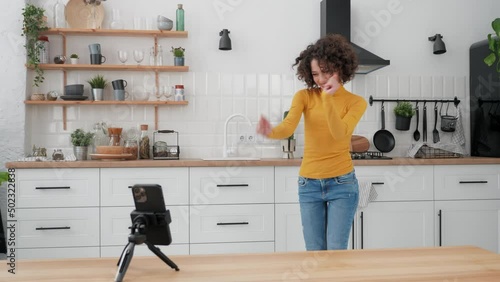 Happy curly woman vlogger dancing on smartphone camera recording video for social media at home kitchen. Smiling positive female blogger influencer listening music records content for social networks photo