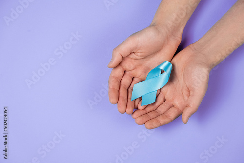 Close up top view of male hands holding small blue awareness ribbon on purple color background wall in studio with copy space for advertisement. Symbol of social and medical issues